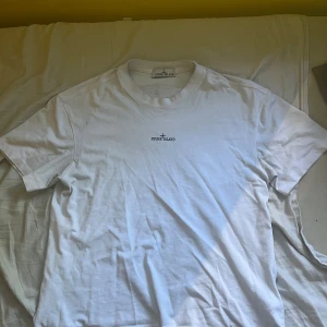 Vit t-shirt från Stone Island - Vit t-shirt från Stone Island med liten logga framtill och stor kompasslogga på ryggen. Klassisk passform och rund halsringning. Tillverkad i mjuk bomull, perfekt för en clean och stilren look.