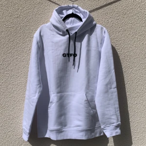 Vit hoodie från GTFO med tryck - Vit hoodie från GTFO med svart tryck framtill och motiv på ryggen. Tröjan har huva med svarta snören och en stor magficka. Tillverkad i bomull och polyester.