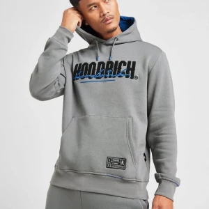 Grå hoodie från Hoodrich - Säljer en grå hoodie från Hoodrich med svart och blå logga på bröstet. Tröjan har en stor magficka, justerbar huva med blått foder och en patch nertill. Perfekt för en avslappnad streetstyle.