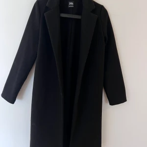 Svart kappa från Zara - Säljer en stilren svart kappa från Zara med klassisk krage och lång passform. Jackan har långa ärmar och enkel design utan synliga knappar eller dragkedja. Perfekt för dig som gillar minimalistisk stil.