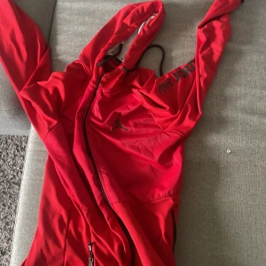 Röd vindjacka/kofta från Air Jordan - Säljer en röd vindjacka från Air Jordan med huva och dragkedja. Jackan har svarta detaljer och logga på bröstet samt texten 'AIR JORDAN' på axeln. Perfekt för dig som gillar sportig stil.