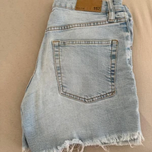 Ljusblå jeansshorts med fransig kant - Säljer ett par ljusblå jeansshorts med rå, fransig kant nertill och klassisk femficksdesign. Shortsen har knappar i gylfen och normal passform. Perfekta för varma dagar och enkel att matcha med allt.