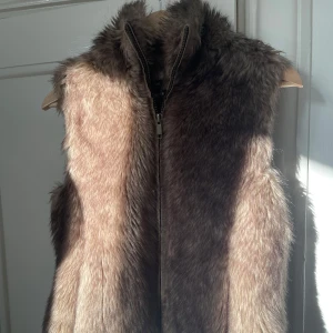 Brun pälsväst med dragkedja - Snygg brun pälsväst med dragkedja framtill. Västen har en naturlig färgskiftning i brunt och beige och är perfekt för att ge din outfit en extra touch. Passar lika bra över en stickad tröja som till en skjorta.
