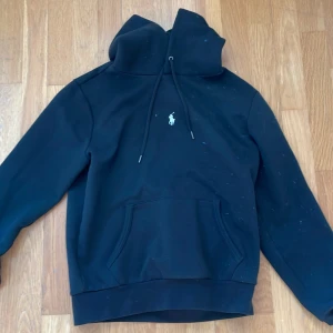 Svart hoodie från Ralph Lauren - Svart hoodie från Ralph Lauren med klassisk logga broderad på bröstet. Tröjan har huva med dragsko och en stor magficka framtill. Perfekt för en avslappnad stil.