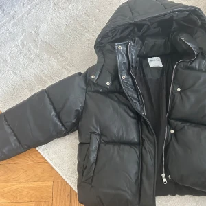 Svart pufferjacka från bershka - Säljer en svart pufferjacka från bershka med huva och dragkedja framtill. Jackan är vadderad och har långa ärmar samt två fickor på sidorna. Perfekt för kyligare dagar.