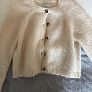 Beige stickad kofta från Atelier - Mysig beige stickad kofta från Atelier med rund hals och stora mönstrade knappar framtill. Koftan har lång ärm och en något croppad passform. Perfekt att slänga över en topp för extra värme. Köpt för 1500