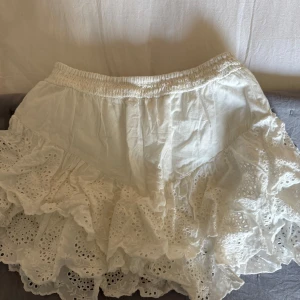 Vit volangkjol med broderade detaljer - Supersöt vit kjol med bred resår i midjan och flera lager volangerna med shorts under. Kjolen har broderade spetsdetaljer och ger en romantisk vibe. Perfekt för varma dagar och lätt att matcha med olika toppar. Endast använd en gång