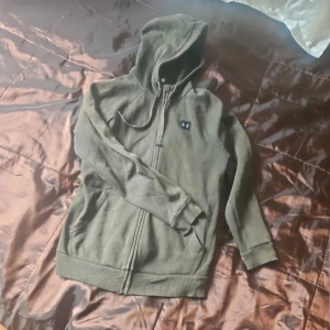 Olivgrön hoodie från Under Armour - Säljer en olivgrön hoodie från Under Armour i storlek S. Tröjan har dragkedja framtill, huva med snörning och två fickor. Perfekt för träning eller chill. Klassisk logga på bröstet och mjukt material. Lagad vid fickan men inget som syns när man bär den.