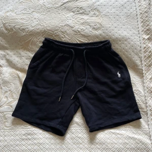 Svarta shorts från Polo Ralph Lauren - Svarta shorts från Polo Ralph Lauren med klassisk broderad logga i vitt på vänster ben. Mjukt material, elastisk midja med snörning och fickor på sidorna samt en bakficka. Perfekta för sommardagar.