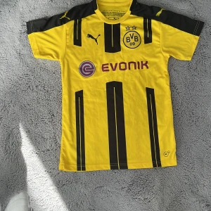 Borussia Dortmund fotbollströja Reus 11 - Gul och svart Borussia Dortmund fotbollströja från Puma med Reus 11 på ryggen. Tröjan har korta ärmar, svarta ränder framtill och klubbmärke på bröstet. Perfekt för fans av BVB och Reus.
