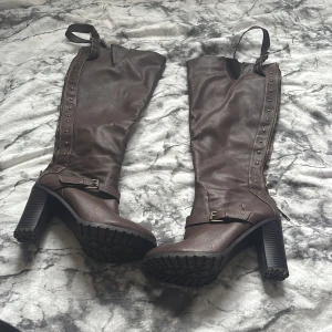 Bruna höga boots med klack och nitar - Säljer ett par bruna höga boots med grov klack och dekorativa nitar längs sidorna. Skorna har dragkedja, spänne och är tillverkade i syntetmaterial. Perfekta för dig som vill sticka ut med en edgy stil.