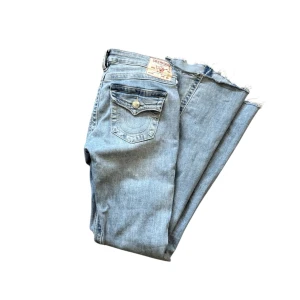 Ljusblå True Religion jeans - Säljer ett par ljusblå jeans från True Religion med flared passform. Lågmidjade och snyggt insydd i midjan. Jeansen har dekorativa bakfickor med lock och knappdetaljer samt klassisk jeanslook. Perfekta för en avslappnad och trendig stil. Midja 35 innerben 75-76