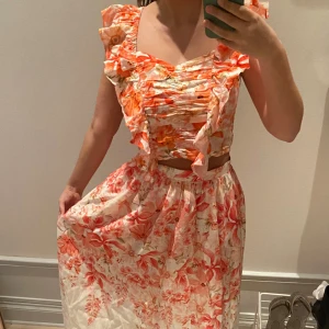 Blommigt set med volang - Superfint set med blommigt mönster i orange och rosa toner på vit botten. Klänningen har volangdetaljer vid axlarna och smockad överdel. Aldrig använt 