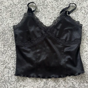 Svart satinlinnetopp med spetsdetaljer - Snygg sexig svart linne-topp i satin med spets längs urringningen och under bysten. Justerbara axelband för perfekt passform. Perfekt att styla till olika outfits.