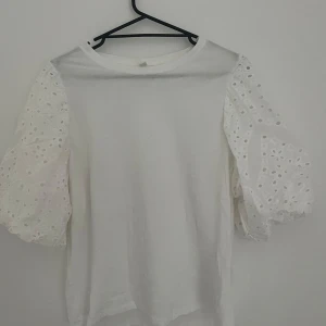 Vit topp med puffärmar och broderade detaljer - Säljer en vit topp med puffiga, broderade ärmar i spetsliknande material. Toppen har rund halsringning och ärmarna har fina hålmönster som ger en romantisk känsla. Perfekt för dig som gillar feminina detaljer!