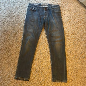 Blå jeans från Jacob Cohën - Snygga blå jeans från Jacob Cohën med klassisk femficksdesign och raka ben. Jeansen har orangea sömmar och en brun patch med logga bak i midjan. Perfekta för en stilren vardagslook.