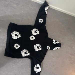Denim tears hoodie  - Säljer en svart hoodie med stora vita blommor tryckta över hela tröjan, även på huvan och ärmarna. Tröjan har en klassisk känguruficka framtill och är perfekt för dig som gillar en unik och trendig stil.