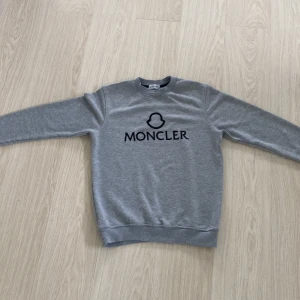 Grå sweatshirt från Moncler - Säljer en stilren grå sweatshirt från Moncler med svart logotyp och tryck på bröstet. Tröjan har rund halsringning, långa ärmar och ribbade muddar vid ärmslut och nederkant. Perfekt för en avslappnad och trendig look.