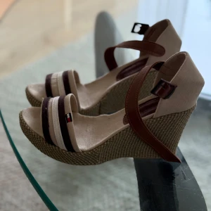 Oanvända beige kilklack sandaler från Tommy Hilfiger - Säljer ett par snygga sandaler med kilklack i storlek 39 från Tommy Hilfiger. Helt oanvända, dock utan prislapp eller kartong. Skorna är beige med bruna detaljer och har en öppen tå samt remmar över foten. Klacken är flätad och ger en somrig känsla. Perfekta för varma dagar!