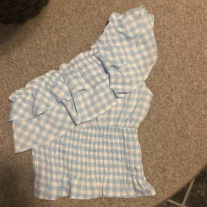 Blåvit rutig one shoulder topp från Boohoo - Säljer en söt blåvit rutig topp från Boohoo med volang och one shoulder-design. Toppen har smockad midja och ett härligt somrigt mönster. Perfekt för dig som gillar unika detaljer och fräsch stil!