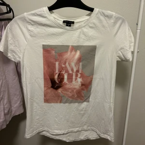 Vit t-shirt med blommigt tryck - Vit t-shirt med ett stort blommigt tryck i rosa. Klassisk rund halsringning och korta ärmar. Perfekt för en avslappnad och trendig look.