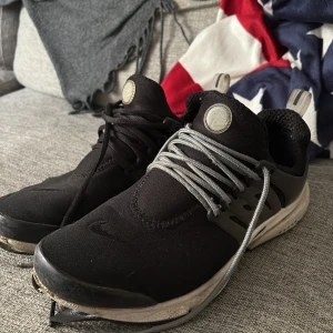 Svarta Nike Presto sneakers - Säljer ett par svarta Nike Presto sneakers med grå snörning och vit sula. Skorna har en sportig design med rund tå och platt sula, samt detaljer i syntetmaterial. Perfekta för träning eller vardagsstil.