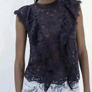 Mörkblå spetsblus med volang - Säljer en mörkblå blus i spets med volangdetaljer vid axlarna. Blusen är ärmlös och har en rund halsringning. Perfekt att styla med jeans eller kjol för en trendig look. Använd ett fåtal gånger.