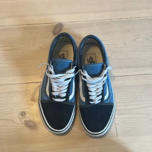 Blå och svarta Vans sneakers - Säljer ett par klassiska Vans sneakers i blått och svart med vit sula och vit snörning. Skorna har den ikoniska vågformen på sidan och en platt gummisula med rutmönster undertill. Perfekta för en avslappnad och stilren look.