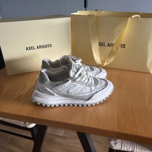 Vita sneakers från Axel Arigato - Säljer ett par snygga vita sneakers från Axel Arigato med chunky sula och coola paneldetaljer. Skorna har snörning och är tillverkade i en mix av skinn och syntetmaterial. Perfekta för dig som gillar stilrena och trendiga sneakers.
