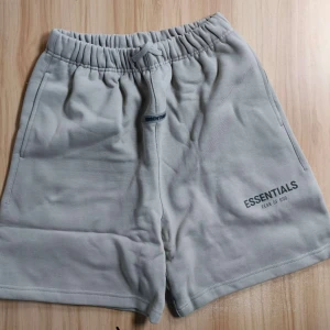 Beige shorts från Essentials - Säljer ett par beige shorts från Essentials med elastisk midja och snörning. Shortsen har fickor på sidorna och en bakficka. Perfekta för en avslappnad stil och har en broderad logga på framsidan.