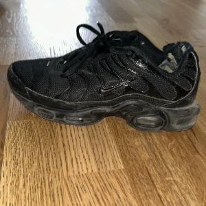 Svarta Nike Air Max Plus TN sneakers - Svarta Nike Air Max Plus TN sneakers med mesh- och syntetöverdel, tydliga TN-detaljer på hälen och vågformade paneler. Skon har snörning och en robust sula med synliga luftkuddar för extra komfort. Perfekt för dig som gillar streetwear.