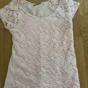 Ljusrosa spetsblus från KappAhl - Söt ljusrosa spetsblusfrån KappAhl i storlek S. Klänningen har blommig spets framtill och på axlarna, med rund halsringning och korta ärmar. Baksidan är i mjuk bomull. 