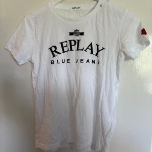 Vit t-shirt från Replay - Säljer en vit t-shirt från Replay med svart text 'REPLAY BLUE JEANS' tryckt på bröstet. Rund halsringning och korta ärmar. Liten röd detalj på vänster ärm. Pris kan diskuteras.