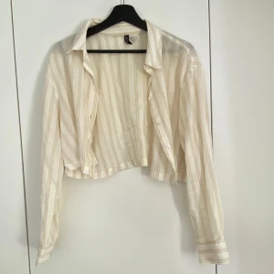 Beige och vit randig croppad skjorta från H&M - Säljer en croppad skjorta från H&M i beige och vitt med tunna ränder. Skjortan har lång ärm, klassisk krage och knappar framtill. 