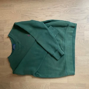 Grön sweatshirt från Polo Ralph Lauren - Mörkgrön sweatshirt från Polo Ralph Lauren med klassisk rund halsringning och ribbade muddar. Tröjan har en liten broderad logga på bröstet och är tillverkad i mjuk bomull. Perfekt för en avslappnad stil. Stl 10-12