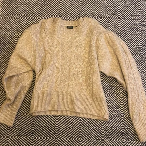 Beige stickad tröja med flätmönster - Säljer en beige stickad tröja från Kappahl med snyggt flätmönster framtill. Tröjan har lång ärm och bred ribbad mudd vid ärmslut och nederkant. Använd en gång men inget tecken på det, nyskick.
