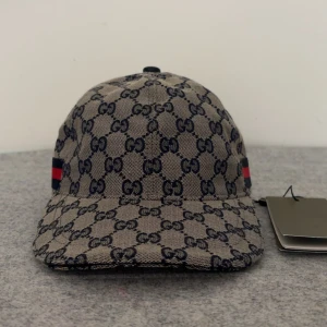 Beige gucci keps - Säljer en beige keps från Gucci med klassiskt GG-monogram i mörkblått och röd-blå rand på sidan. Justerbar rem baktill i skinn. Perfekt accessoar för att lyfta din stil. Storlek ”M”