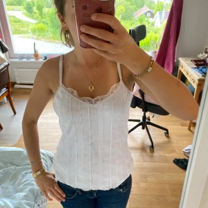 Vit spetsdetaljerad linnetopp - Säljer en söt vit linnetopp med tunna axelband och spetsdetaljer längs urringningen. Toppen har dekorativa sömmar och en feminin, romantisk känsla. Perfekt att matcha med jeans eller kjol.