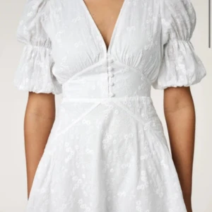 Vit blommig omlottklänning med puffärm - Superfin vit klänning med blommigt broderat mönster, v-ringning och knappar framtill. Klänningen har puffiga korta ärmar och markerad midja för en feminin siluett. Perfekt för sommarens alla tillfällen!