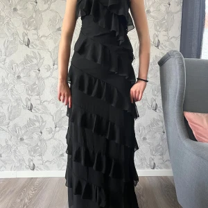 Svart långklänning med volanger - Säljer en svart långklänning med flera lager av volanger som ger en cool och elegant look. Klänningen har smala axelband och en figurnära passform upptill. Perfekt för dig som vill sticka ut på festen! Priset kan diskuteras vid intresse!!