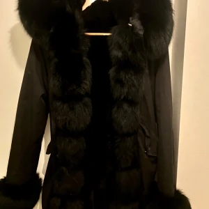 Svart parkajacka med äkta päls  - Säljer en svart parkajacka med stor, fluffig päls längs huva, krage och ärmslut. Jackan har lång ärm och en klassisk parkamodell med markerad midja. Perfekt för kyliga dagar.