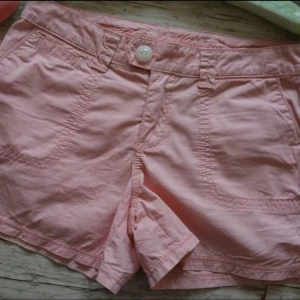 Low waisted shorts - Säljer ett par ljusrosa shorts med en låg midja. Shortsen har två framfickor, två bakfickor med lock och knapp, samt en knapp i midjan. Perfekta för varma dagar och enkel att matcha med olika toppar.