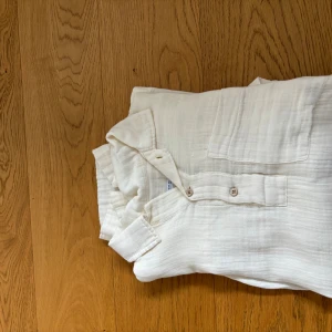 Vit kortärmad henley i bomull - Säljer en stilren vit henley-skjorta i mjuk bomull med korta ärmar. Skjortan har knappar framtill och en bröstficka som ger en chill och avslappnad vibe. Perfekt för varmare dagar när du vill ha något luftigt och bekvämt.