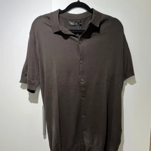 Brun kortärmad skjorta - dressman - Stilren brun skjorta från dressman med korta ärmar och klassisk krage. Skjortan har knäppning framtill med matchande knappar och är tillverkad i ett mjukt material som känns skönt mot huden. Perfekt för en avslappnad och snygg look.