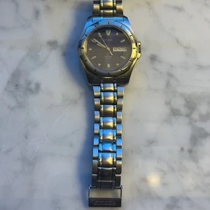 Citizen herrklocka i silver - Stilren herrklocka från Citizen med rund svart urtavla och dag- och datumvisning. Klockan har ett klassiskt armband i silverfärgad metall och tydliga indexmarkeringar. Perfekt accessoar för dig som gillar tidlös design.