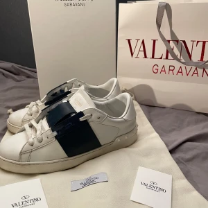 Valentino open - Säljer ett par vita Valentino Garavani Open sneakers med bred mörkblå rem över mitten. Skorna har rund tå, snörning och platt sula. Klassisk och stilren design med exklusiv känsla. Perfekta för dig som gillar lyxiga sneakers.