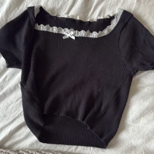 Svart ribbad topp med spets och rosett - Supersöt svart ribbad topp med korta ärmar, vit spetskant och en liten rosett framtill. Toppen har en croppad passform och är perfekt för dig som gillar en gullig och trendig stil. Materialet är stretchigt och bekvämt.