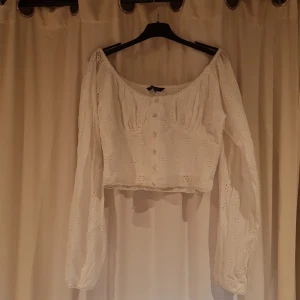 Vit blus med broderade blommor Amelie & Me - Superfin vit blus från Amelie & Me med broderade blommor och croppad passform. Blusen har långa puffiga ärmar, knappar framtill och smockad rygg. Perfekt för dig som gillar romantisk och söt stil med offshoulder-ringning. Kontakta för fler bilder eller frågor.