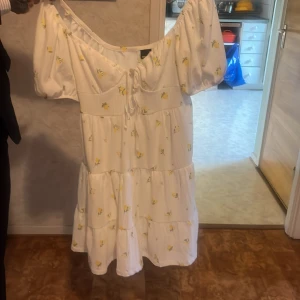 Vit blommig kortklänning Gina Tricot - Superfin vit kortklänning från Gina Tricot med små gula blommor, puffärmar och snörning framtill. Klänningen har en luftig passform och är perfekt för varma dagar och har en söt, romantisk vibe.