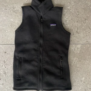 Svart fleeceväst från Patagonia - Svart fleeceväst från Patagonia med hög krage och hel dragkedja framtill. Västen har två fickor med dragkedja och en liten broderad Patagonia-logga på bröstet. Perfekt till sommaren och våren. Passar XS